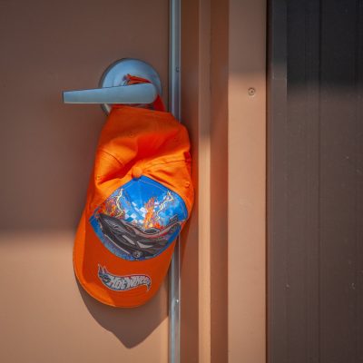 A close-up shot of a bright orange baseball cap with a Hot Wheels logo and car design hanging from a silver door handle.