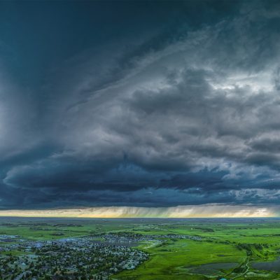 Wide aerial panorama of an immense, dark storm cloud formation hanging low over bright green fields and a distant town.