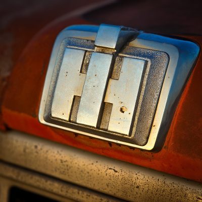Extreme close-up of a weathered chrome IH logo on a rusted orange truck hood.