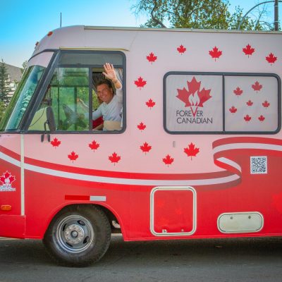 A pink and red bus decorated with maple leaves, with a man waving from the driver's window.
