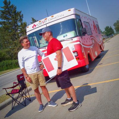 Two men stand on an asphalt lot in front of a white and red motorhome featuring "Forever Canadian" text. One man is in a white shirt and khaki shorts, while the other wears a red shirt and baseball cap, holding a plastic Canadian flag sign.