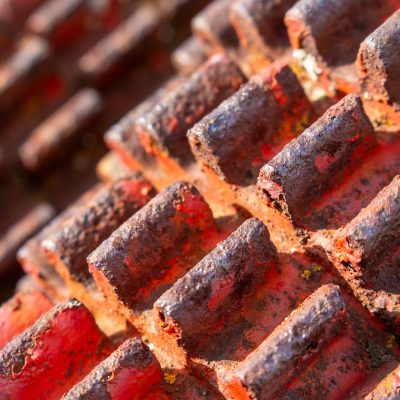 A close-up, shallow depth-of-field shot of rusted red metal gear teeth on agricultural machinery, highlighting the flaking paint and textured brown rust in warm sunlight.