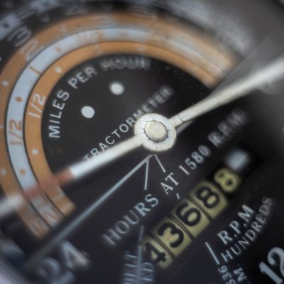 Extreme close-up of a vintage tractor meter gauge. The black dial shows speeds, hours, and engine RPM with weathered needles and an odometer.