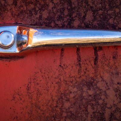 Close-up of a shiny chrome vintage car door handle on a rusty, weathered red metal surface.