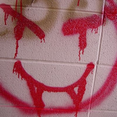A close-up of a light beige concrete block wall featuring a crude, dripping red spray-painted smiley face with crossed-out eyes and a wide grin.