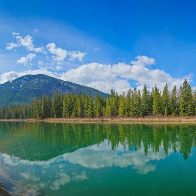 A wide landscape view of a calm, turquoise river reflecting a clear blue sky, white clouds, a dark mountain, and a row of green evergreen trees. A walking path is visible on the left bank.