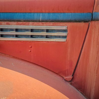 Close-up of the textured side panel of a rusty red vintage car with a blue trim line and silver chrome vents.