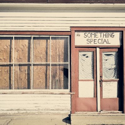 A straight-on shot of an abandoned wooden building with dirty white siding and dark red trim. A large window on the left is boarded up with plywood, while the door on the right has a sign above it reading "SOMETHING SPECIAL". The glass windowpanes of the double door are covered from the inside with torn, faded white paper.