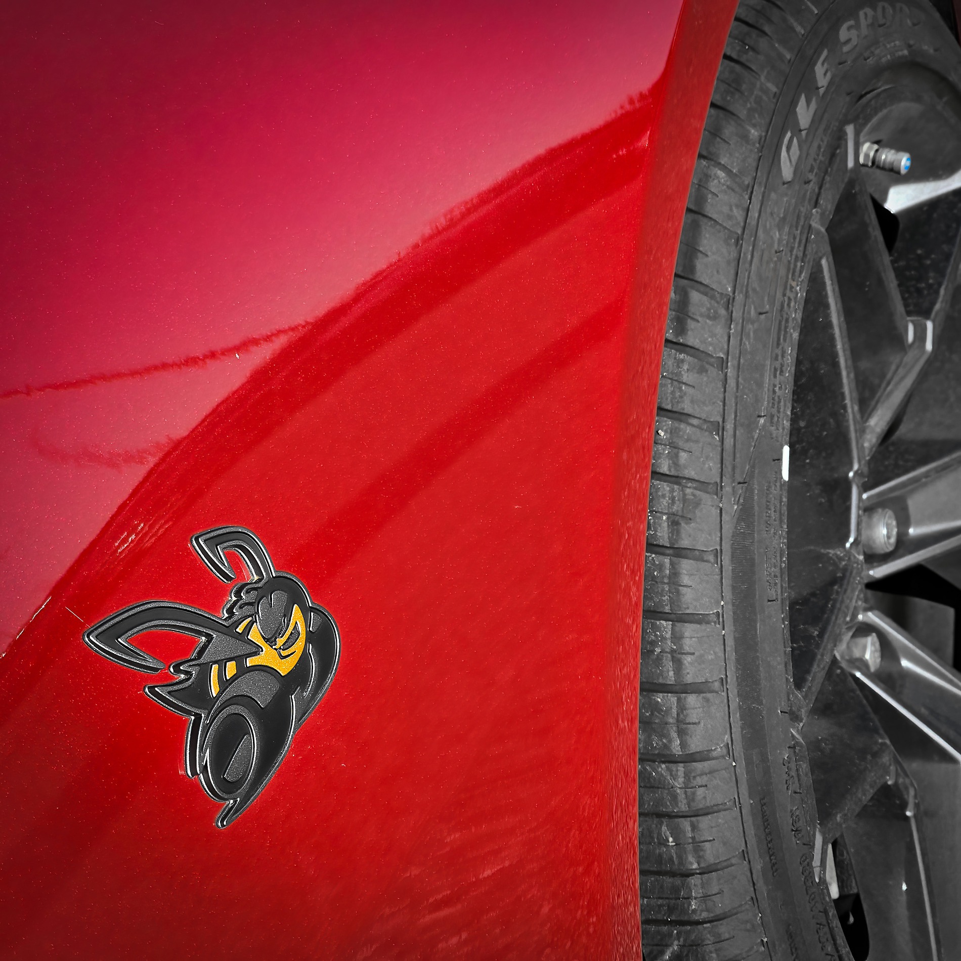 Detailed close-up of a glossy cherry red car fender featuring a black and yellow angry bee mascot emblem, positioned next to a black multi-spoke wheel and tire.
