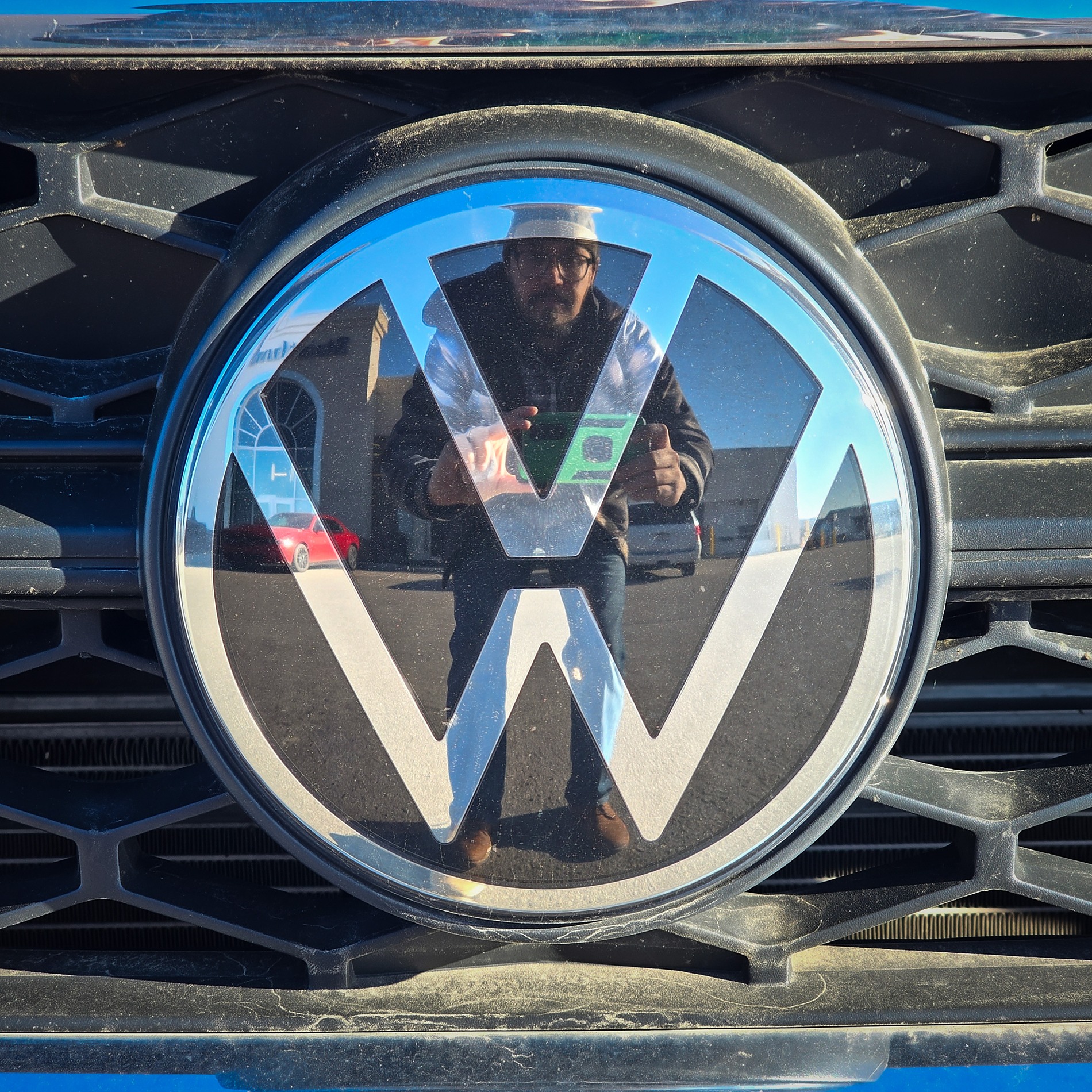 A close-up of a circular chrome Volkswagen logo set against a black honeycomb grille. The reflective surface of the emblem shows a clear mirror image of a man wearing glasses and a cap, holding up a smartphone to take a photo. In the background of the reflection, a red car and a dealership building are visible.