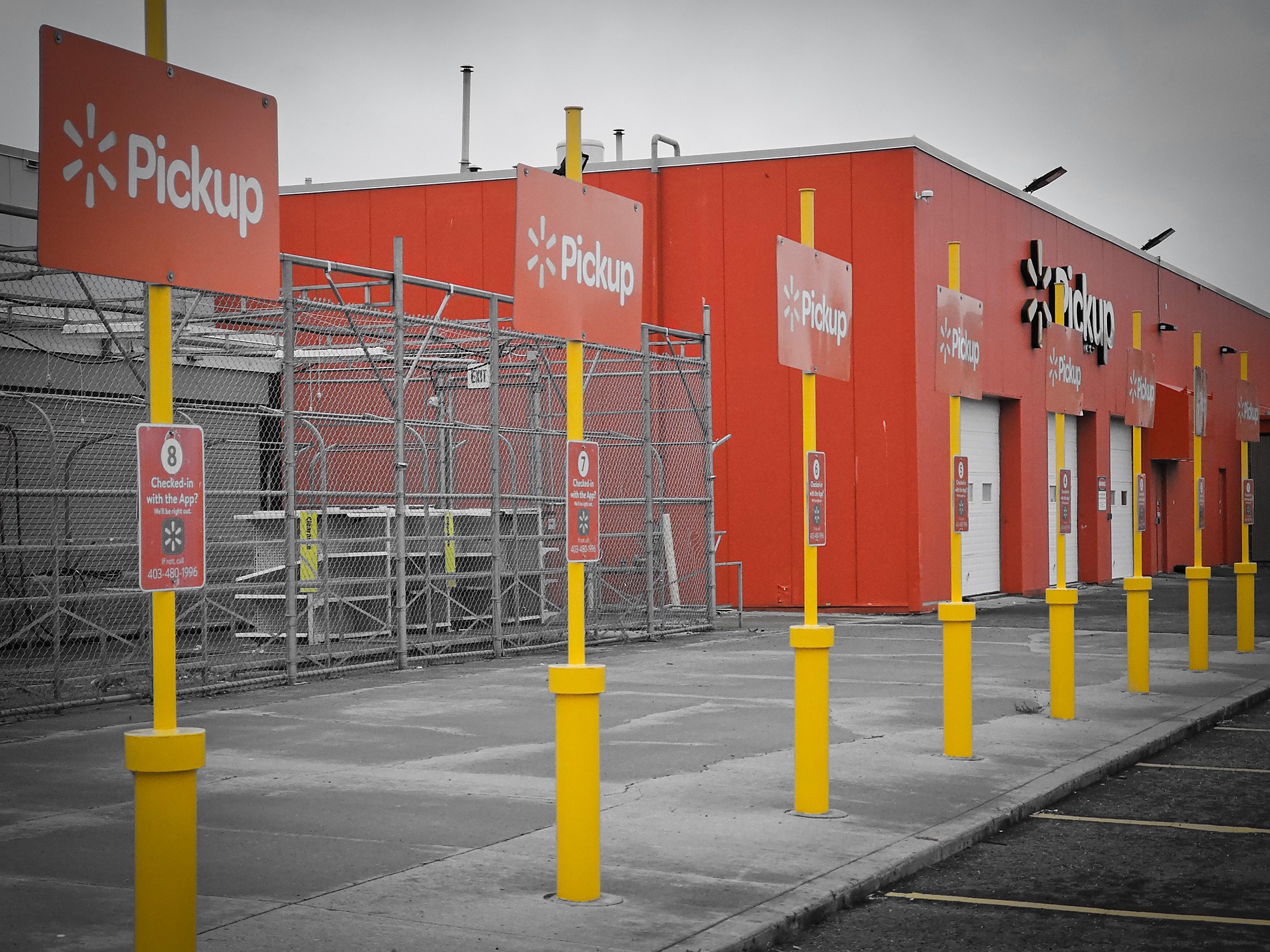 A perspective shot of multiple yellow posts with orange Walmart Pickup signs in a parking lot, next to a large orange store building, with selective coloring emphasizing the yellows and oranges against a desaturated grey background.