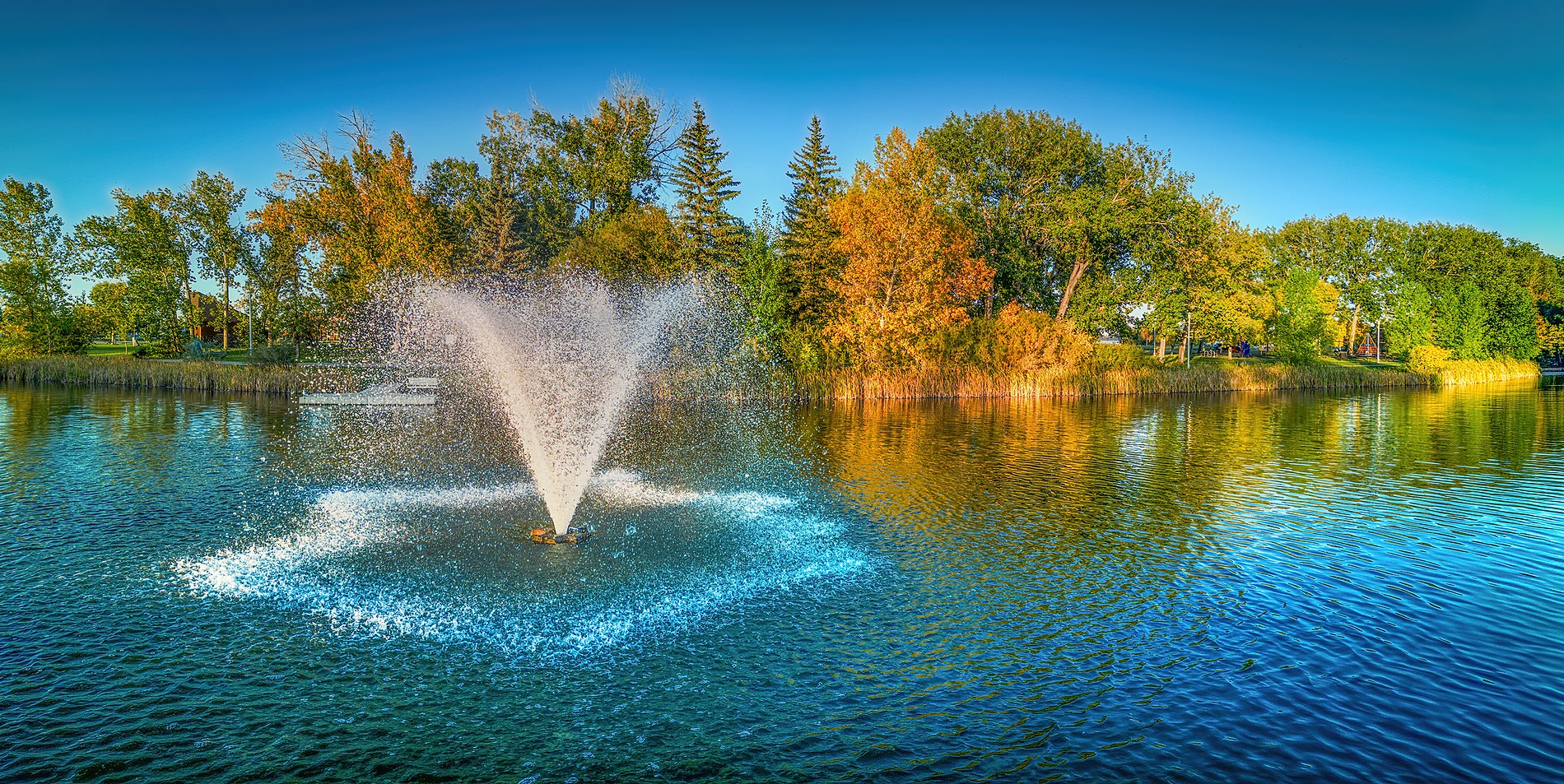 A wide panoramic shot of a water fountain spraying in a symmetrical plume on a calm lake, with green and yellow autumn trees lining the shore under a clear blue sky.