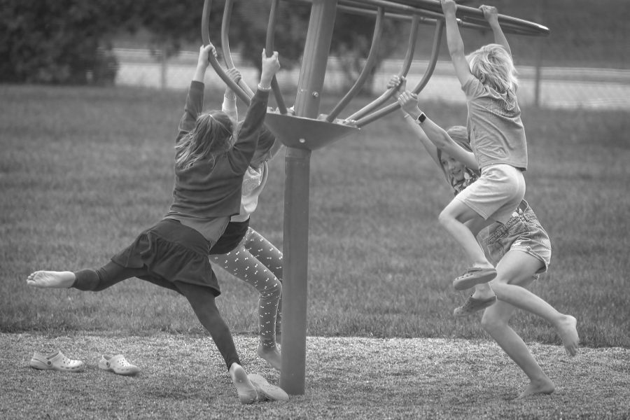 A black and white photograph showing four children holding onto the metal ring of a playground spinner carousel, suspended mid-spin. They hang in various dynamic poses over a gravel pit, with a grassy field in the background.