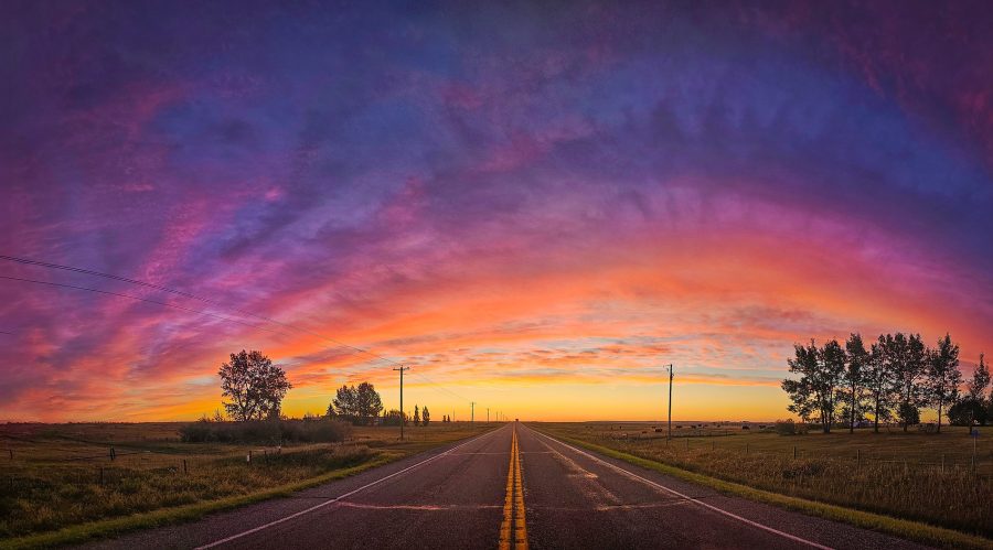 A wide-angle panoramic view of an empty two-lane asphalt road with a double yellow line stretching straight toward a vibrant sunrise. The vast sky above is filled with sweeping clouds transitioning from deep purple and magenta to bright orange and yellow near the horizon, casting a warm glow over the flat rural landscape.