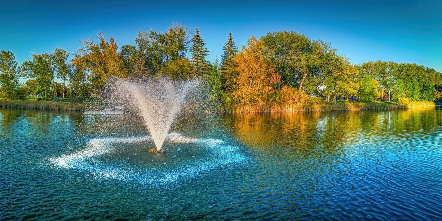 A wide panoramic shot of a water fountain spraying in a symmetrical plume on a calm lake, with green and yellow autumn trees lining the shore under a clear blue sky.
