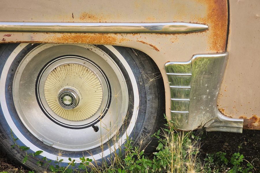 Detailed close-up of a vintage Dodge wheel well featuring a whitewall tire, ribbed cream hubcap, rusted tan fender, and chrome trim standing over green grass.